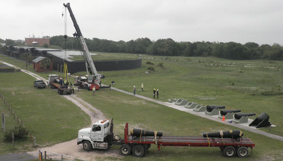 Large cannons are lifted by heavy crane and placed on flatbed trucks.