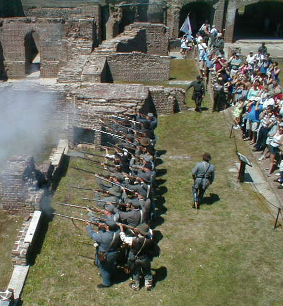 A line of Civil War reenactors fire muskets while visitors watch.