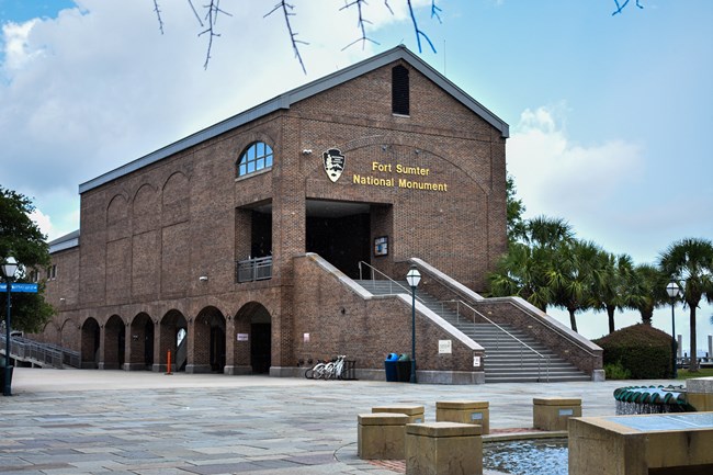 A large brick building with a golden National Park Service logo on the front. The entrance is accessible via stairs leading up to a second story landing.
