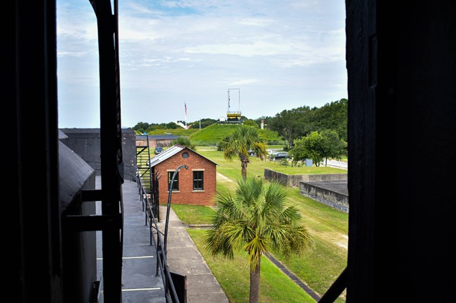 A view from Battery Jasper. A brick storehouse is in the foreground. In the background, the American flag is visible as well as a yellow building called the Harbor Entrance Command Post.