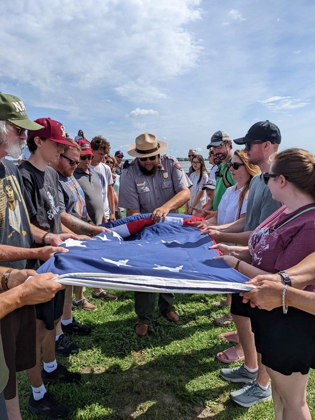 A Ranger unfolding a very large American flag with the help of visitors wearing tee shirts, hats, and general tourist clothing