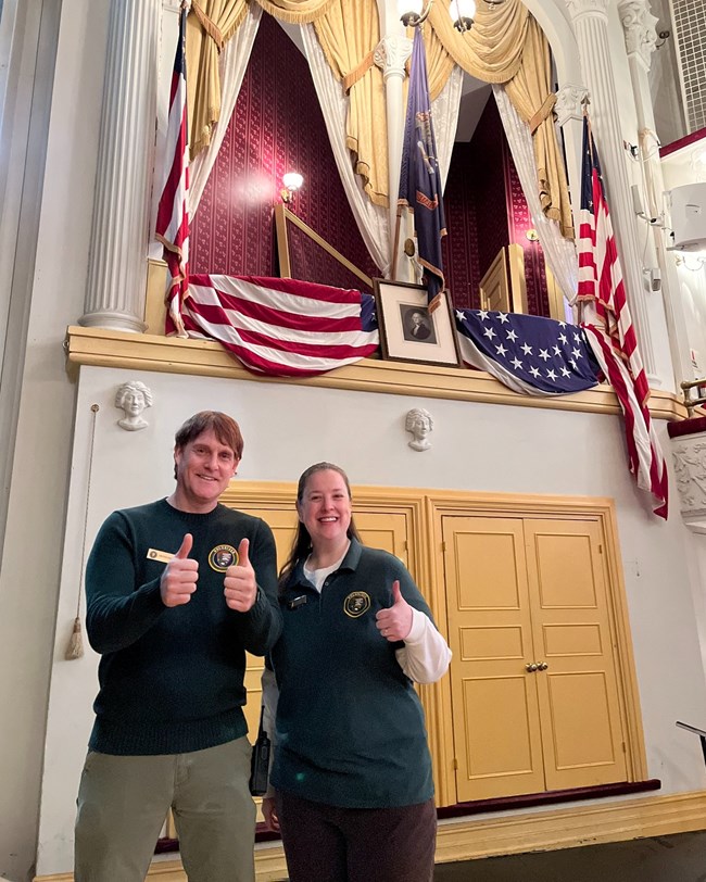 A man and a woman wearing green NPS volunteer polo shirts stand on the Ford's Theatre stage below Abraham Lincoln's state box