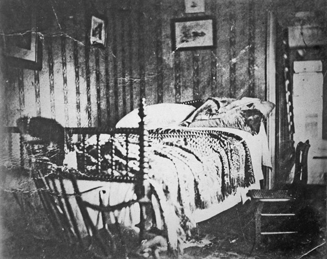 A bedroom with pictures hanging on the wall and patterned wallpaper. Two wooden chairs are visible. The spindle bed in the center of the image is disheveled from use, including a bloody pillow.