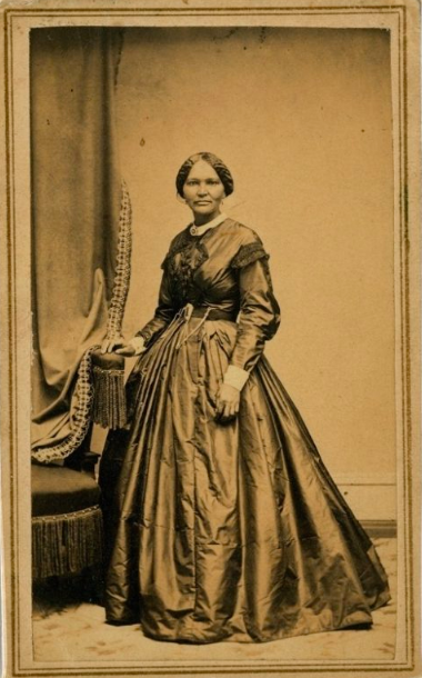 Black and white photo of an African American woman wearing a full length 19th century-style dress and standing with her right hand resting on her chair