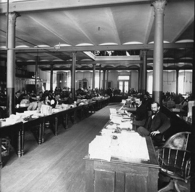 black and white photo of a large room with dozens of men sitting and working at tables.
