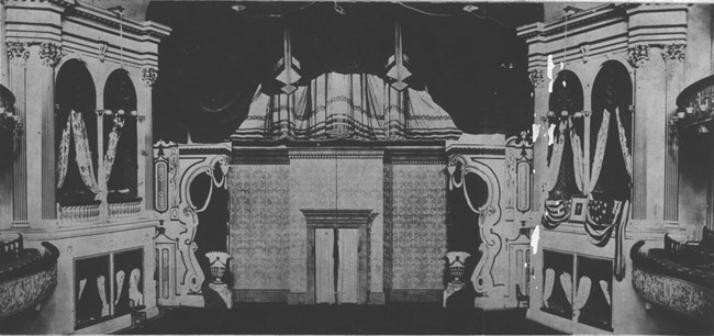 Photograph of the interior of Ford's Theatre, 1865. The theatre stage is decorated with curtains and an elaborate home interior set piece. The theatre box in which President Lincoln sat is adorned with American flags and a portrait of Washington.