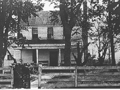 Black and white photo of a modest two story wood frame farmhouse with a porch across the front