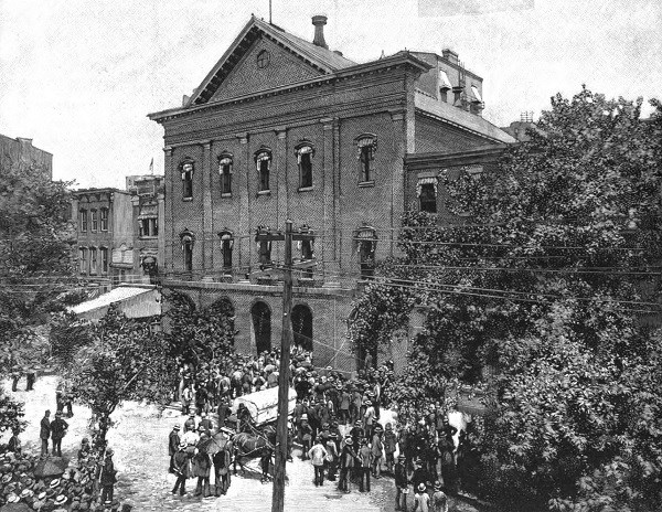 Large three story building with 5 bays, windows in each story and bay, and arched entrances. A large crowd and wagon is gathered around the entrances.