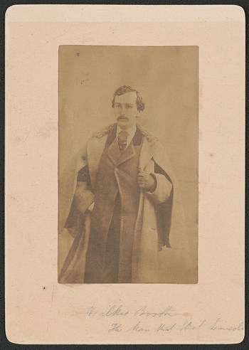 3/4 length portrait photo of John Wilkes Booth wearing a suit, tie, and heavy overcoat. The text on the photo matte reads "Wilkes Booth The Man that Shot Lincoln."
