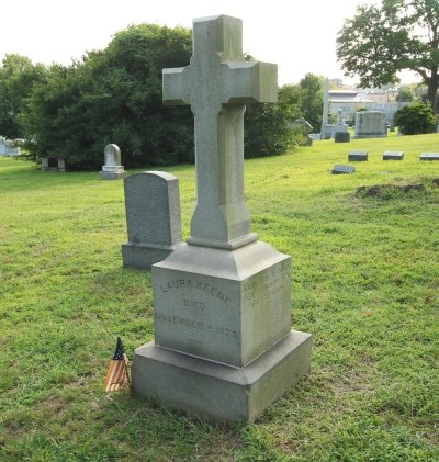 Granite cross-shaped gravestone on a square base and inscribed with Laura Keene's name, in a grassy area of a cemetery