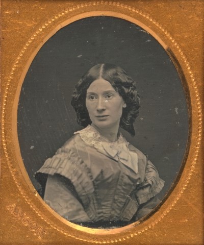 Circular Black and White photo in a rectangular gilt frame depicting a white woman with dark hair with curls dressed in a fancy blouse with lace accessories.