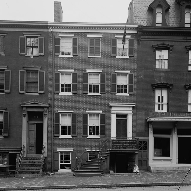A four-story house with three windows on the upper two levels and two windows on the lower two levels. A sign identifies the house as containing the Oldroyd Lincoln Memorial Collection. A small child sits near a pillar at the base of the house.