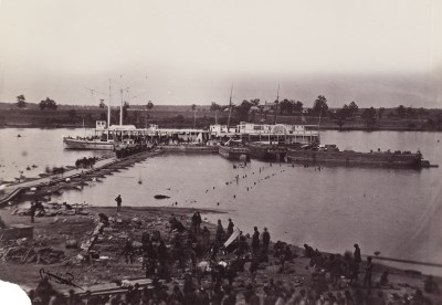Black and white 1860s photo of barges moored at a dock in the middle of a river.