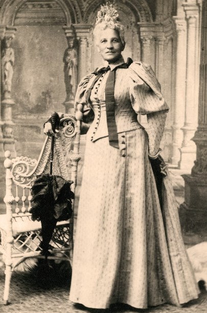 Black and white photo of an older African American woman standing with her right arm resting on a chair and holding a closed parasol