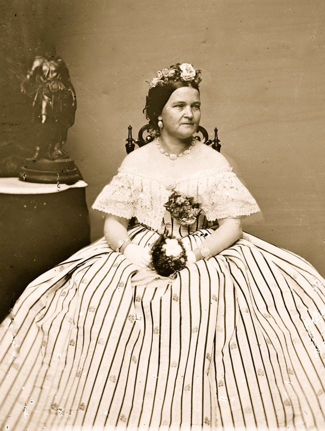 Sepia tone photo of a seated woman (Mary Lincoln) wearing a striped dress with a lace blouse top accented by flowers. She also wears flowers in her hair.