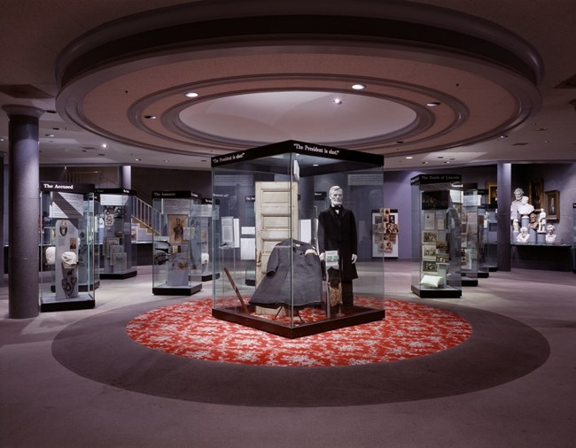 Photo of Fords Theatre Museum with large central circular carpet and floor to ceiling display case with Lincoln figure displaying his clothing and personal effects.