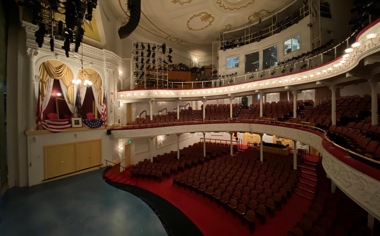 Photo of theatre stage and audience seating on three levels, with the box seats decorated with flags on the other side of the stage