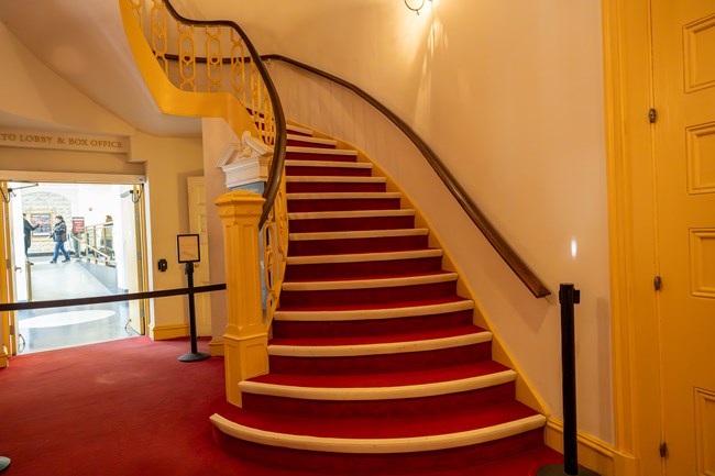 A carpeted staircase curves up above a doorway blocked off by a retractible belt.