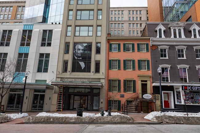 A row of conjoined buildings of different heights and building materials on a street, one of which has a portrait of Lincoln in the window next to a shorter brick building with a staircase up to the front door.