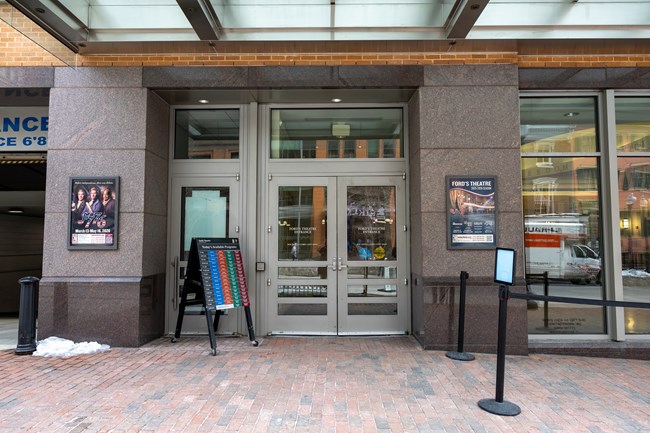 Glass double doors at the front of a building with a glass overhang and a wide brick sidewalk out front.
