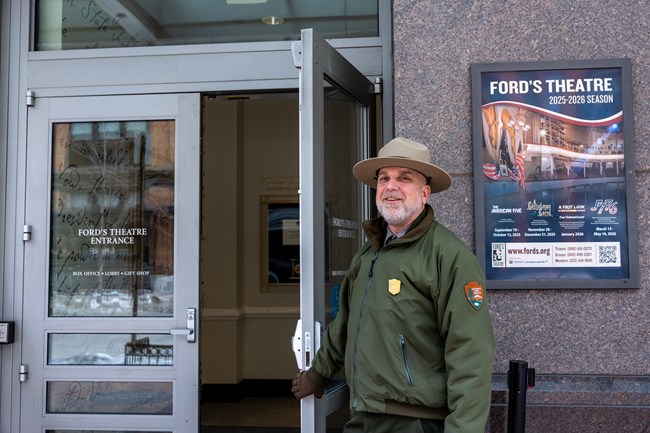 A smiling man in a green jacket and a tan hat with a round brim holds open a glass door.