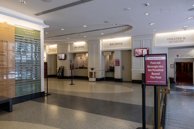 A large room with a bench next to the wall, three counters with windows in the middle with box office signs over each one, and a wide ramp down towards doors to a theatre.