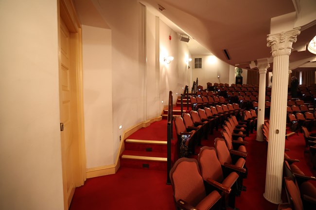 Red carpeted steps with a handrailing lead along a wall with rows of tiered theatre seating to the right.