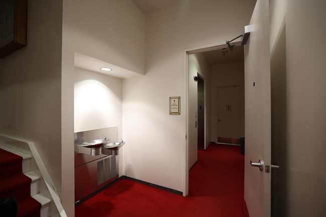 Two drinking fountains at different heights next to a doorway to a hallway with an elevator and a door to a men's restroom.