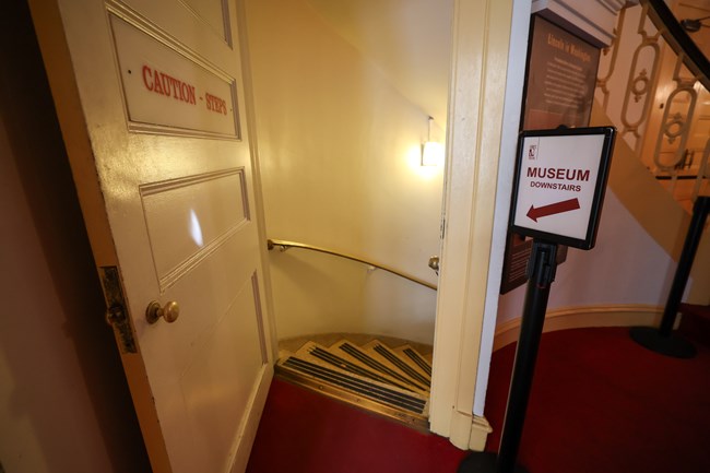 An open door to a staircase leading down with a sign for a museum next to the doorframe.