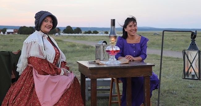 two women dressed in 19th century dresses sitting in front of oil lamp