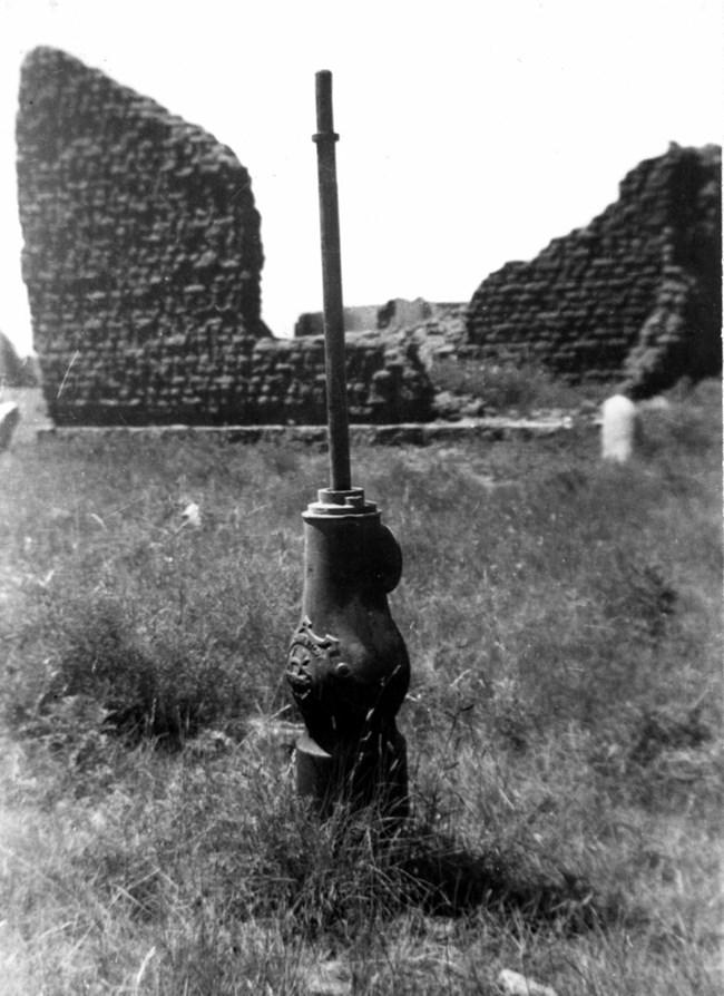 Late 1800s fire hydrant standing amid fort ruins. An embossed metal base sits close to the ground, while a metal pole extends upward twice the length of the base.