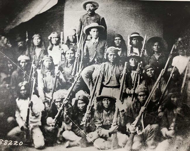 Group of seated Indian warriors holding rifles with army officer in middle