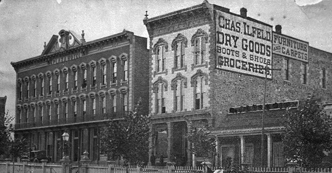 Two large brick buildings side by side with sign on one side that says "Chas. Ilfeld Dry Goods Boots and Shoes Groceries"