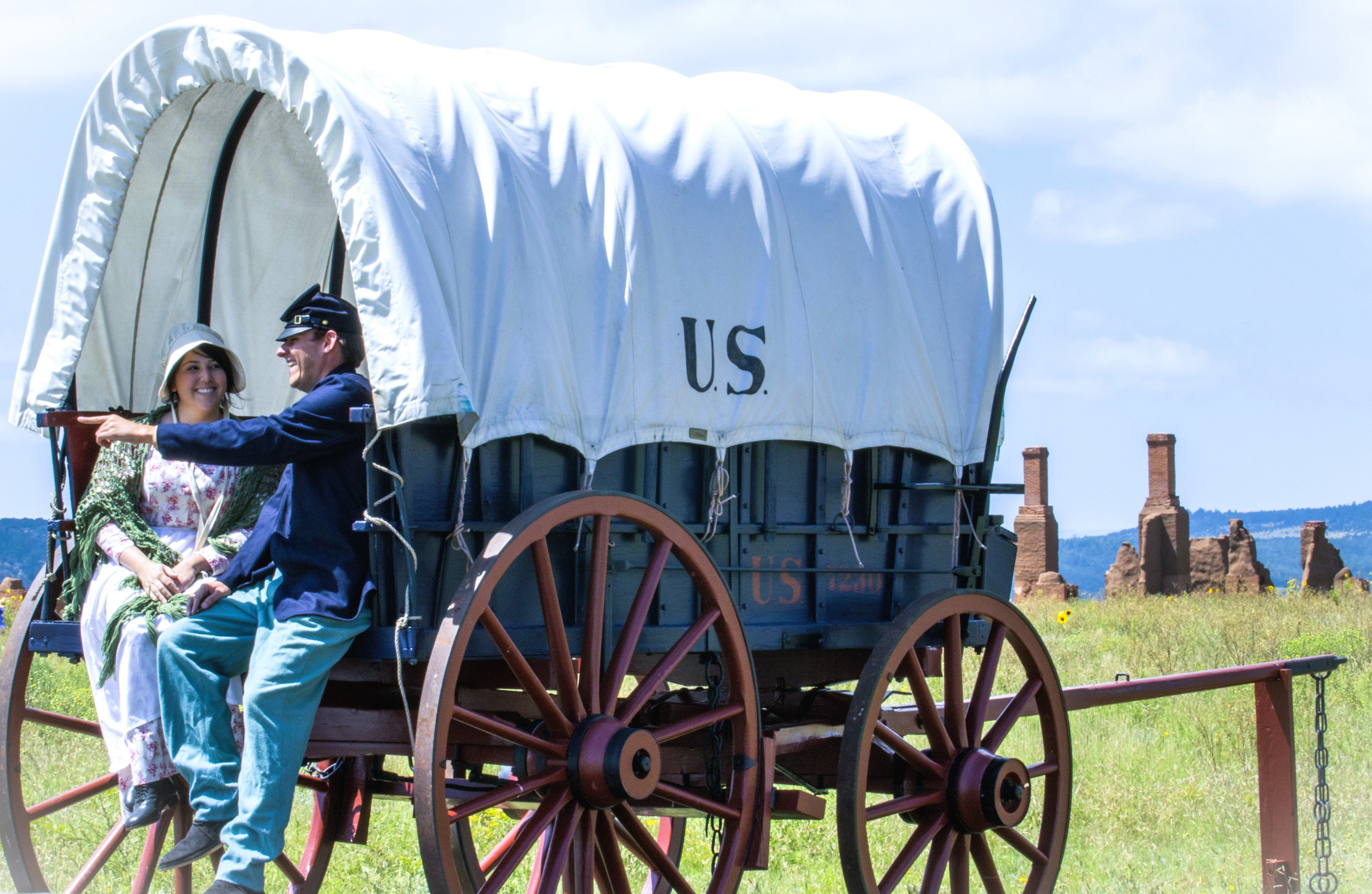 Fort Union ruins and covered wagon. Two people sitting in a covered wagon in 1800s style clothing with adobe ruins in the background.