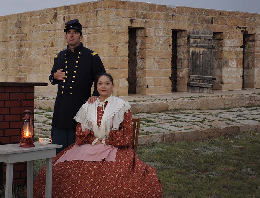 Army officer standing beside his wife, who is seated next to table with tea cup