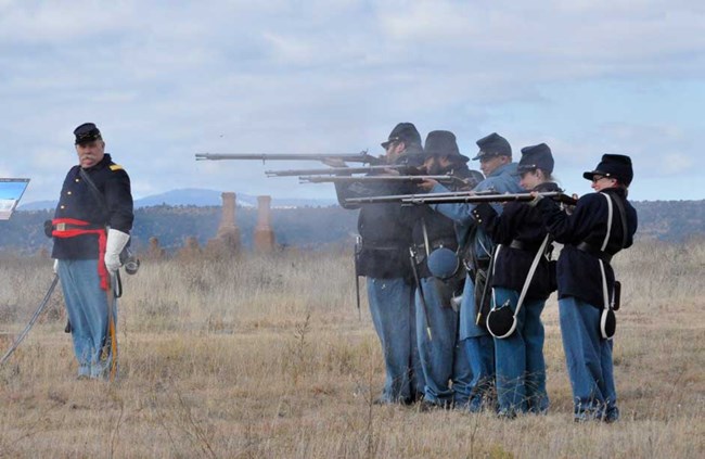 A group of people dressed as Union Civil War soldiers standing shoulder to shoulder in line holding rifles extended in front of them with smoke emerging from muzzles of weapons