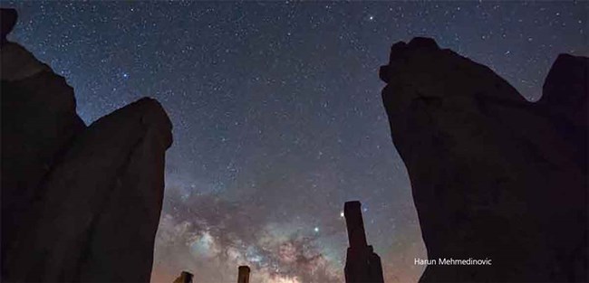 A dark sky full of stars with the Milky Way at one side and adobe walls silhouetted in front of sky.