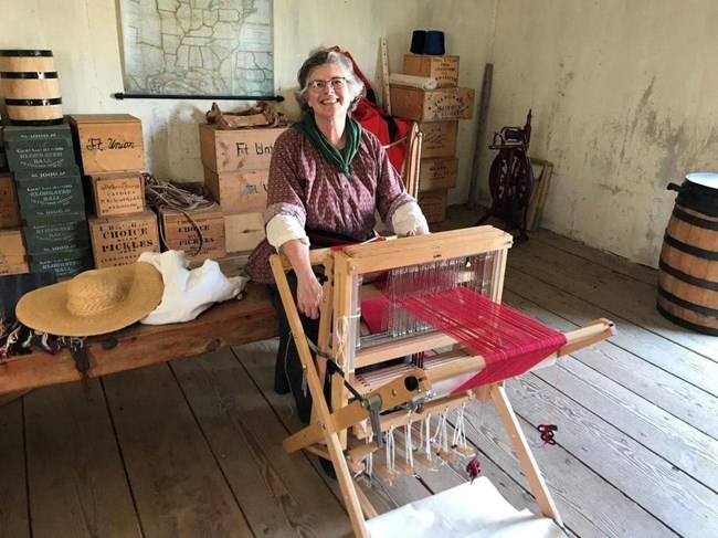 Woman in old fashioned dress sits at a weaving loom, bright red thread is wrapped around the loom..