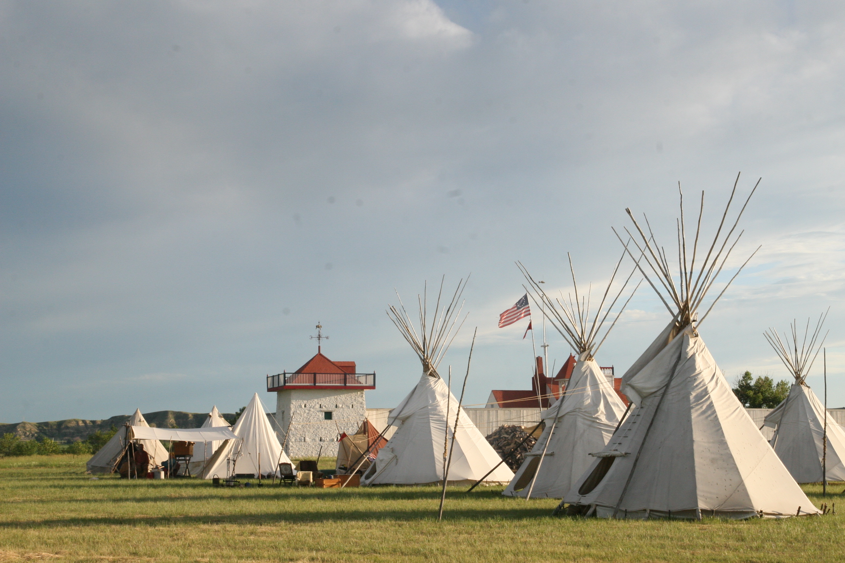 Rendezvous Camp Three tall tipis stretch into a cloudy blue sky, other tents are visible behind the tipis, and a stone bastion or square blockhouse topped with a red roof is visible in the far center.