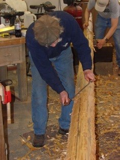 Men using hand tools to smooth the round surface of a falgpole in a workshop.