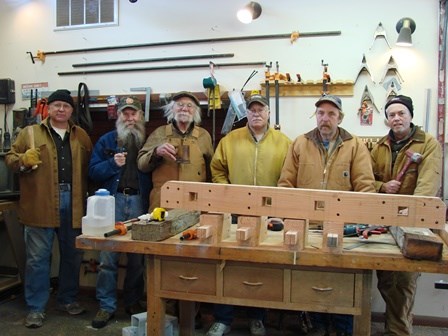 Six men pose in woodworking shop with tools behind a piece of the flagpole with tools on the wall behind them.