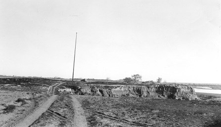 Black and white photo of dirt road through a field leading to a flagpole with a river below.