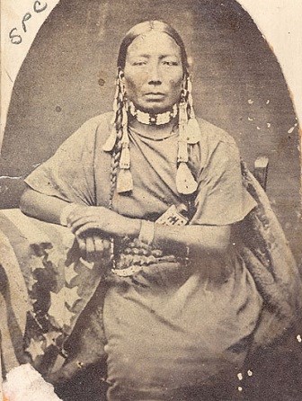 Black and white photo of American Indian woman sitting. She wears a solid cloth dress, a beaded choker, and two bracelets. Her hair is braided with shells.