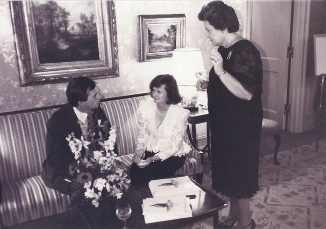 Black and white photo of a man sitting on a couch talking to a woman sitting next to him and a woman standing beside them.