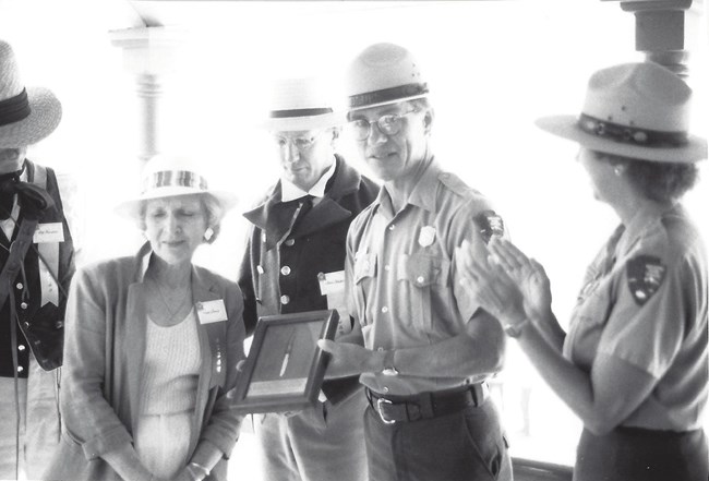 Black and white photo of a Park Ranger holding display box containing a pen, three people looking on.