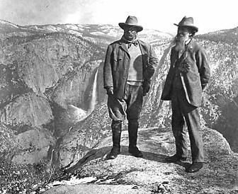 Black and white photo of two men posed on rock outcrop, mountain valley visible behind them.