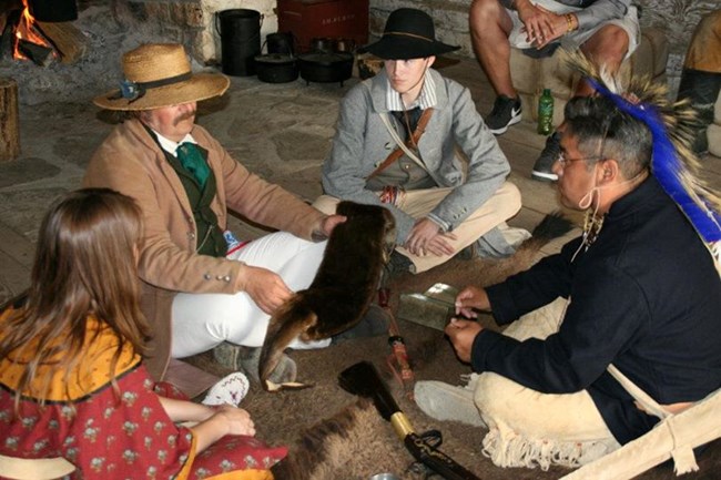 Four people in period clothing sit in a circle. One man holds a brown fur for another to examine.