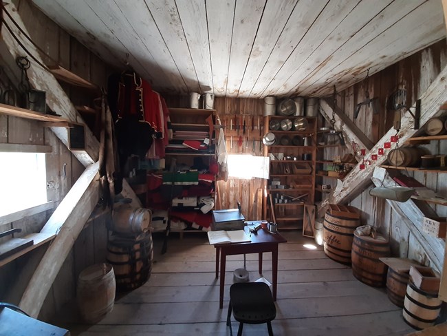 Square room with rustic wooden shelves filled from floor to ceiling with historic items like blankets, knives, tin cups and beads strands, and large barrels on the floor. There is a small desk with a chair. On two walls there are small square windows.