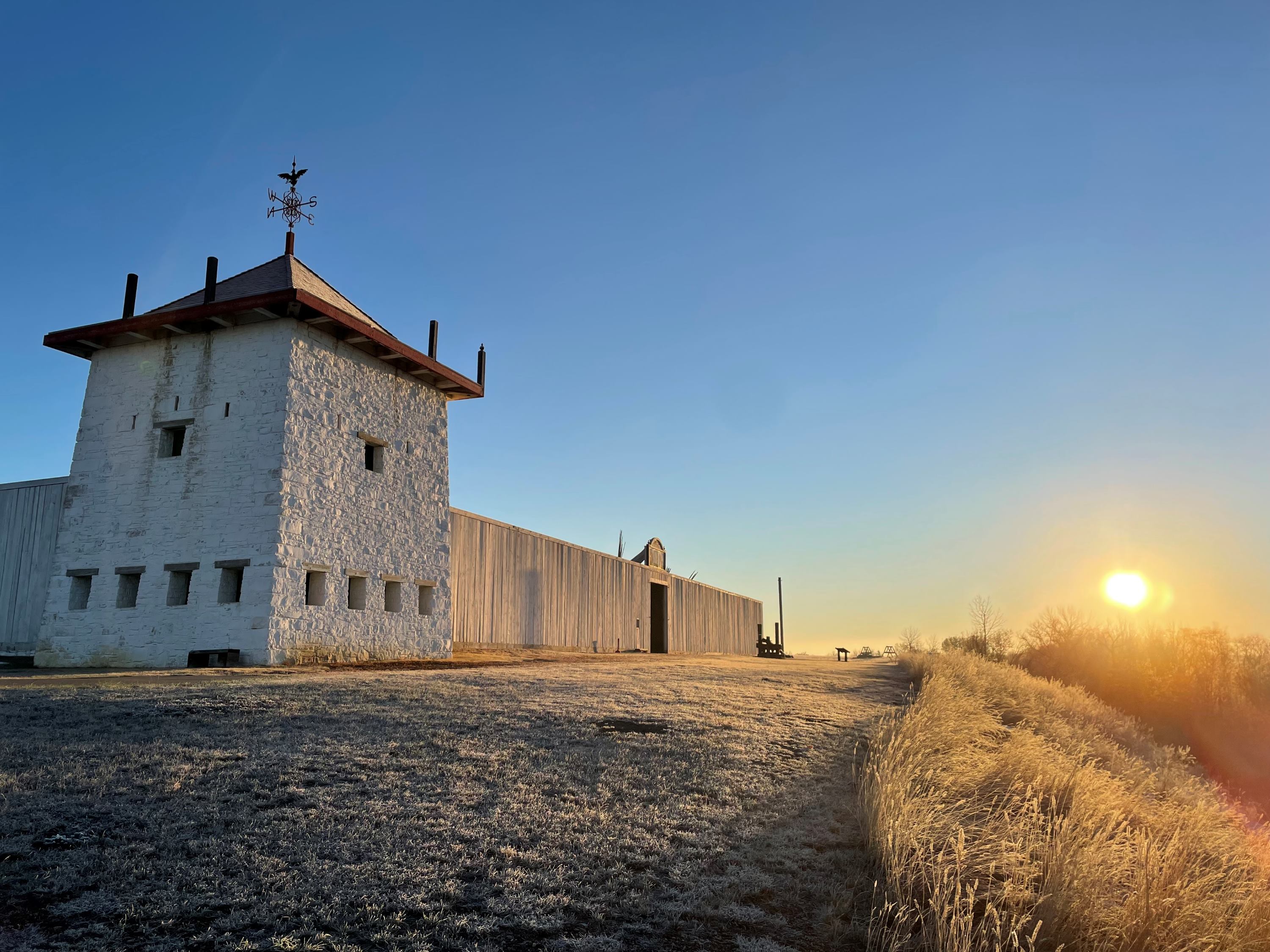 Sun glistens off of the snow-covered ground, large white stone bastion and white wooden walled fort stretches up towards a clear blue sky.