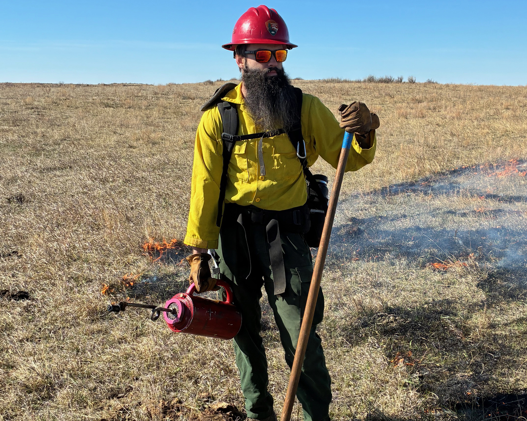 A firefighter wearing green pants, yellow shirt, hard hat and carrying tools puts fire on the landscape.